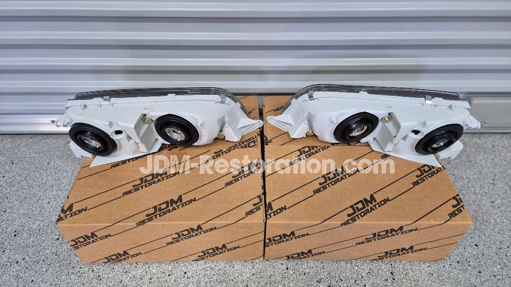Car headlights on a cardboard box with JDM Restoration.com branding against a metallic background.