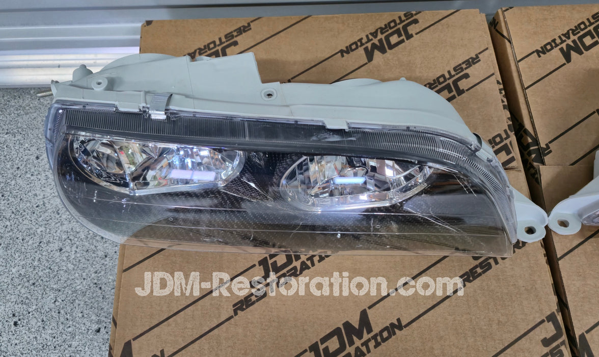 Car headlights on a cardboard box with JDM Restoration.com branding against a metallic background.
