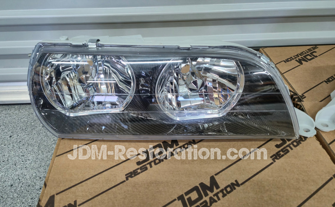 Car headlights on a cardboard box with JDM Restoration.com branding against a metallic background.