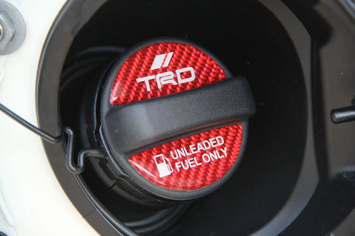 A close-up of a black and red carbon-fiber-patterned fuel cap with white TRD and 'UNLEADED FUEL ONLY' text, set inside a dark circular opening.