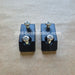 A pair of black 3D-printed rectangular blocks with two silver screws each, placed on a brown textured surface. A watermark 'JDM- Restoration.