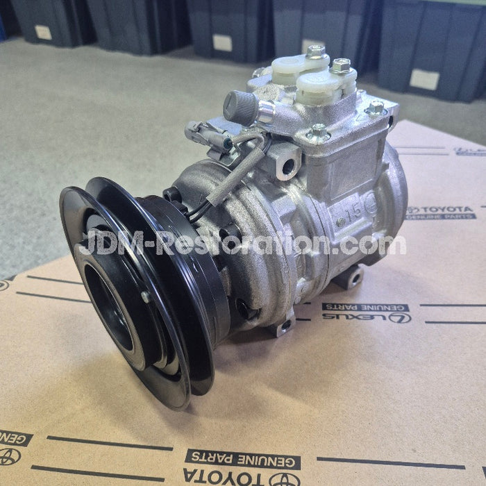 A used silver automotive air conditioning compressor with a black pulley, resting on a cardboard box printed with Toyota Genuine Parts branding.