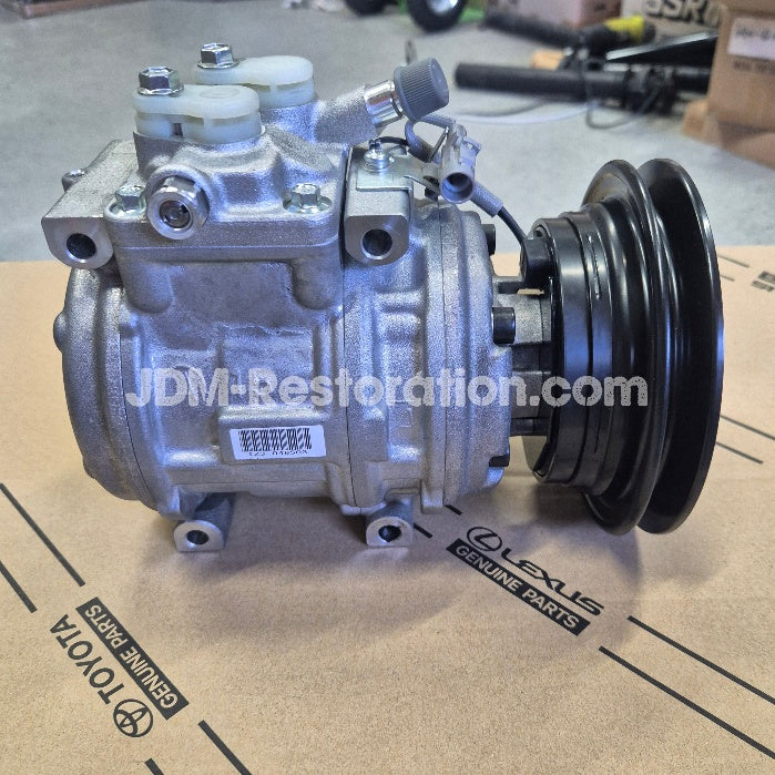 A silver and black automotive air conditioning compressor pulley assembly resting on a cardboard box printed with Lexus and Toyota branding. A translucent JDM Restoration watermark is visible across the center.