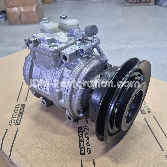A used automotive air conditioning compressor assembly with a black pulley, photographed on a cardboard box printed with Toyota and Lexus Genuine Parts logos.