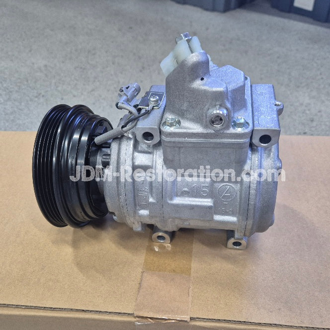 A silver automotive air conditioning compressor unit with a black pulley belt and a white plastic connector, resting on a cardboard box. A watermark 'JDM-Restoration.
