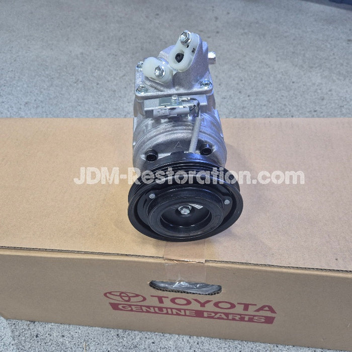 A used silver and black automotive air conditioning compressor assembly resting on a cardboard box with a visible Toyota Genuine Parts logo. A watermark for 'JDM-Restoration.