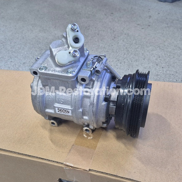 An automotive air conditioning compressor pulley assembly with a silver metal body and a black ribbed pulley, resting on a cardboard box with a concrete floor in the background.