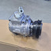 An automotive air conditioning compressor pulley assembly with a silver metal body and a black ribbed pulley, resting on a cardboard box with a concrete floor in the background.