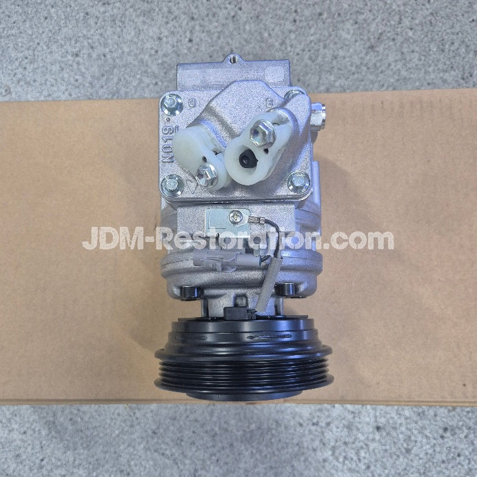A silver metal automotive air conditioning compressor pulley assembly with a black ribbed belt pulley and white plastic connectors, photographed on a cardboard box over a gray concrete floor. A watermark 'JDM-Restoration.