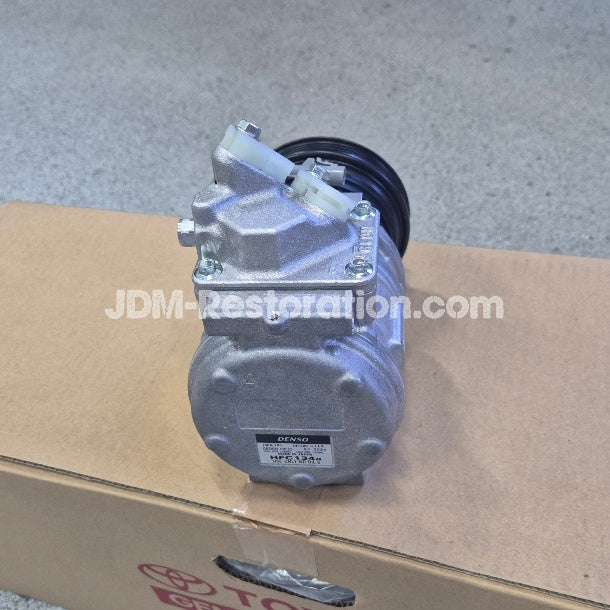 A silver metallic automotive pump or compressor unit with a black hose attached, standing on top of a brown cardboard box. A white JDM-Restoration.