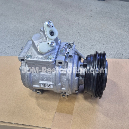 Silver automotive air conditioning compressor pulley assembly with a black ribbed pulley belt, photographed on a cardboard box with a concrete floor background.