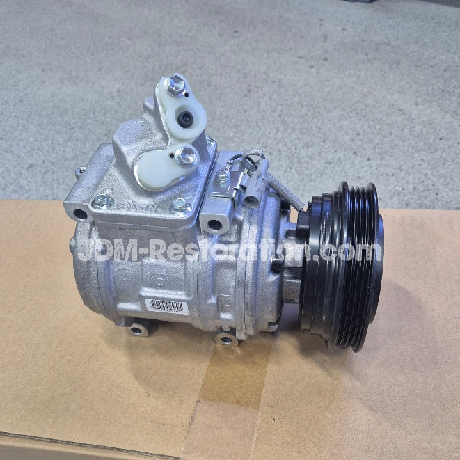 Silver automotive air conditioning compressor pulley assembly with a black ribbed pulley belt, photographed on a cardboard box with a concrete floor background.