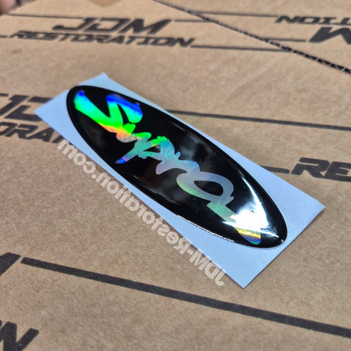 A glossy black oval decal sticker with holographic rainbow lettering, shown on its light blue backing sheet over a brown cardboard box.