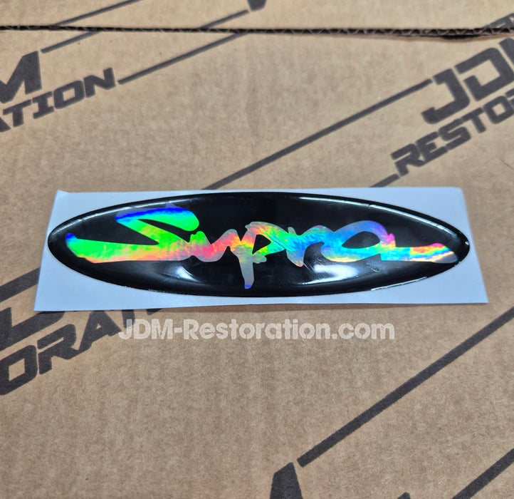 An oval holographic vinyl decal featuring the word 'Supra' in a stylized script, shown on its light blue backing sheet against a cardboard background with JDM Restoration branding.