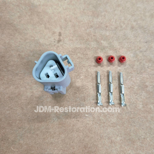 A light gray electrical connector with three red rubber boots and three metal terminal leads laid out on a tan textured surface. A JDM-Restoration.