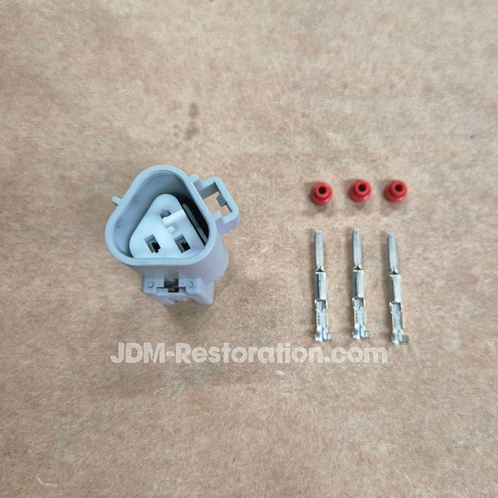 A light gray electrical connector with three red rubber boots and three metal terminal leads laid out on a tan textured surface. A JDM-Restoration.
